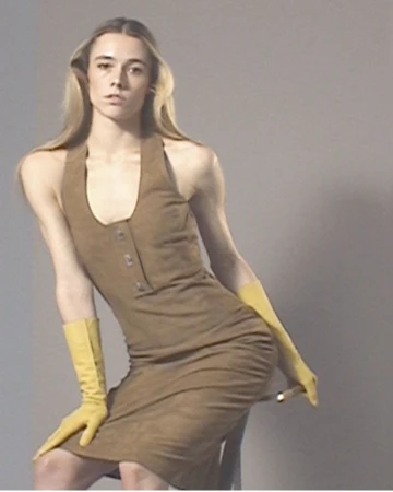 A person with long blonde hair, wearing a brown halter dress and yellow gloves, poses in a studio setting with a grey backdrop.