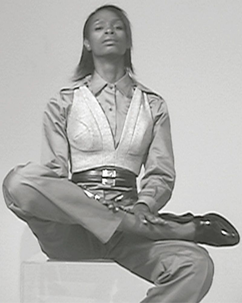 A slim African-American woman poses indoors against a plain background wearing a cropped top, collared shirt, slacks, and loafers.