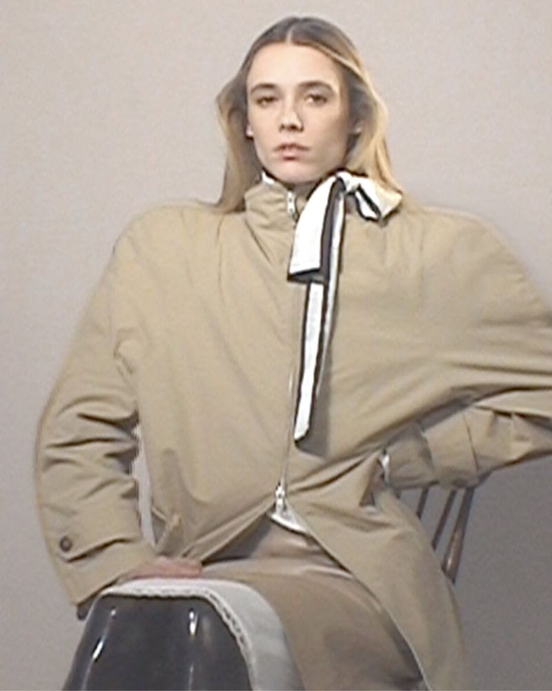 Young adult female model in a beige trench coat poses sitting on a chair in a studio with a plain white background.