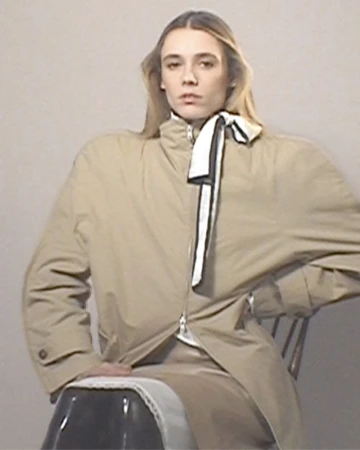 Young adult female model in a beige trench coat poses sitting on a chair in a studio with a plain white background.