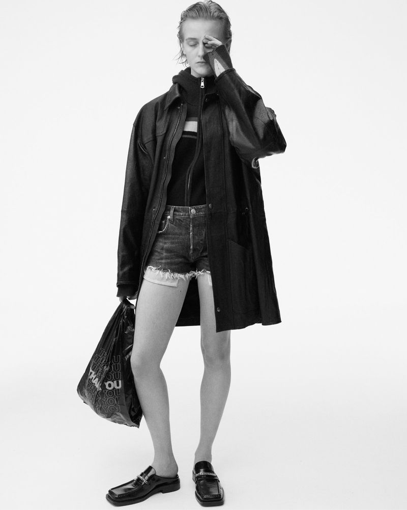 Black and white studio shot of a fashion model in denim shorts and oversized coat holding a shopping bag. The model is covering their face.