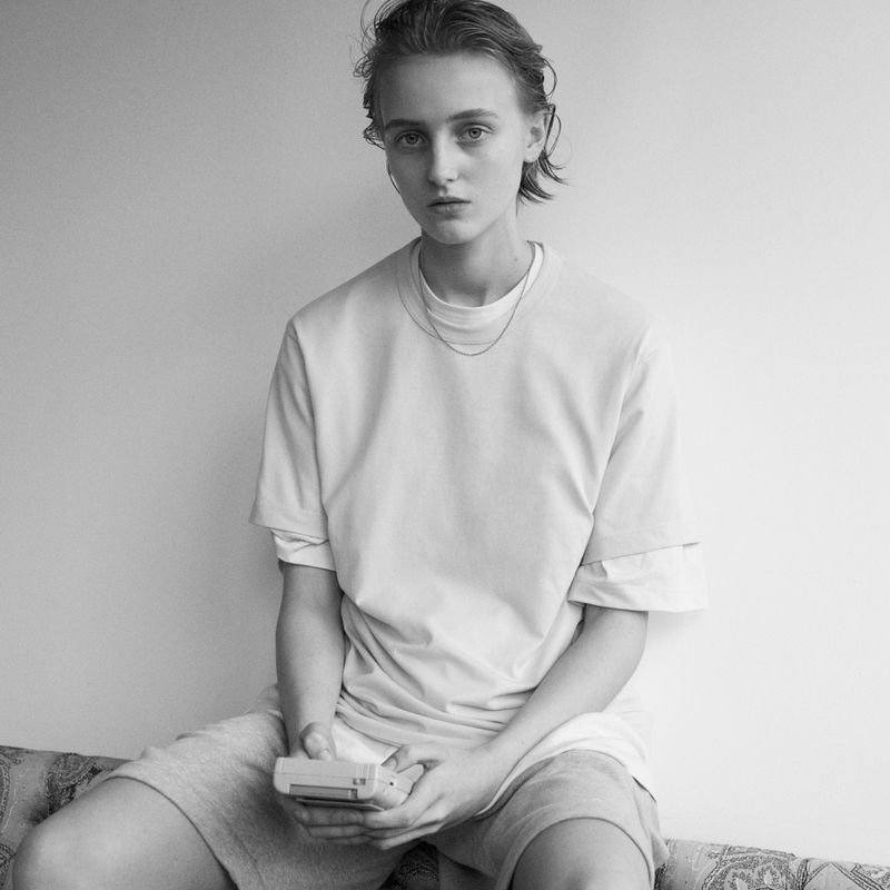 Black and white portrait of a young adult with short hair, wearing a t-shirt and shorts, sitting on a patterned couch, holding a handheld device.