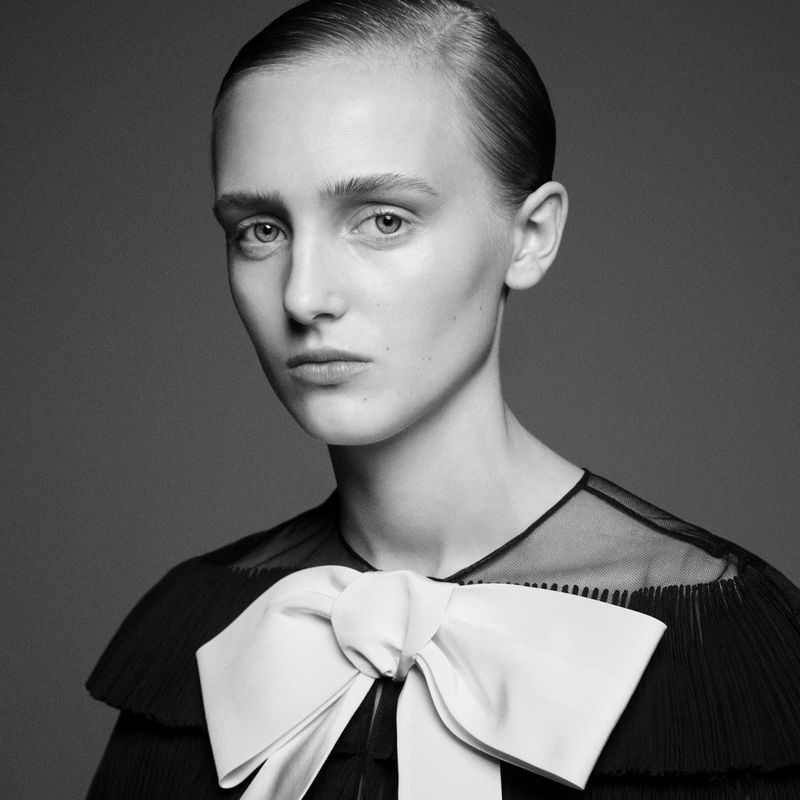 Black and white portrait of a woman with slicked back hair wearing a dark dress with a large white bow, captured in a photo studio.