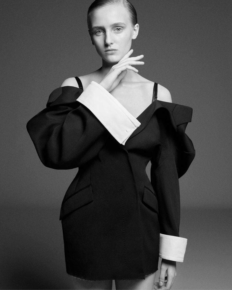 Black and white portrait of a woman with slicked-back hair wearing an oversized black blazer as a dress in a neutral studio.