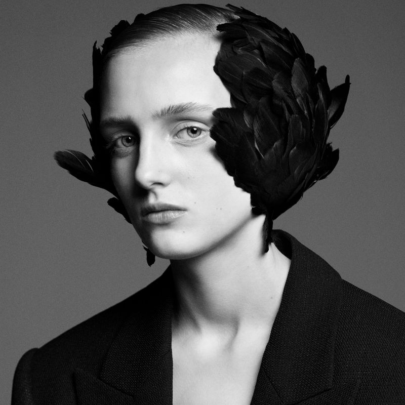 Portrait of woman with black feathers adorning head, wearing a blazer against a gray background in a studio setting.