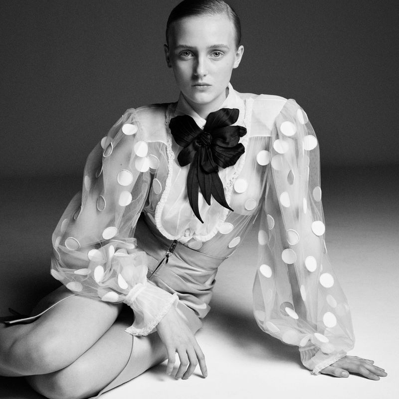 Monochrome studio portrait of a woman with slicked-back hair, wearing a sheer polka-dot blouse with a large black bow, sitting on the floor.