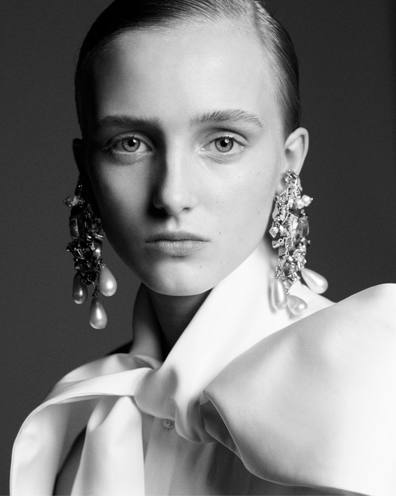 Black and white fashion portrait of a fair-skinned woman with statement earrings and a large white bow blouse against a grey backdrop.