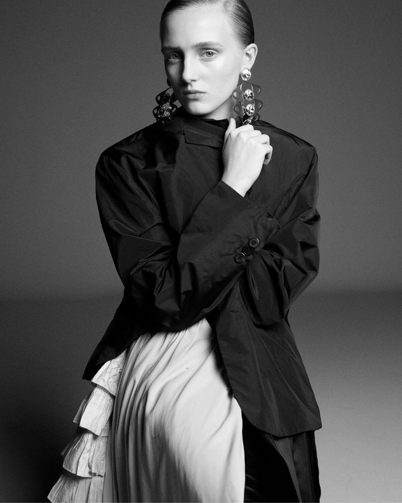 Black and white fashion portrait of a fair-skinned young woman in an oversized black jacket and white skirt with ruffle details.