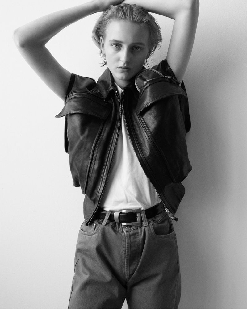 Monochrome portrait of a young adult with slicked-back hair, wearing a leather vest, t-shirt, jeans, and belt against a plain wall.