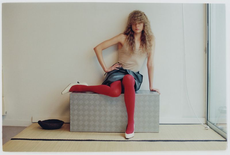 A young woman poses indoors, seated on a metal box with red tights and white heels, against a neutral wall and a glimpse of a window.