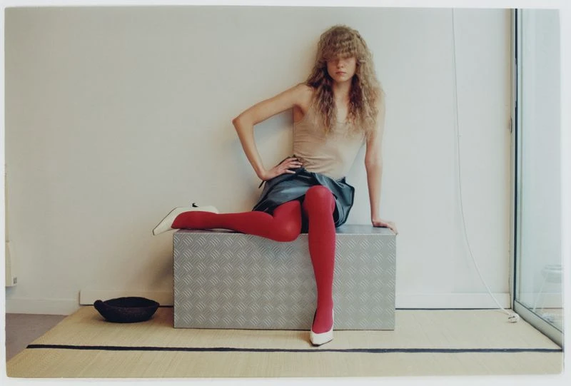 A young woman poses indoors, seated on a metal box with red tights and white heels, against a neutral wall and a glimpse of a window.