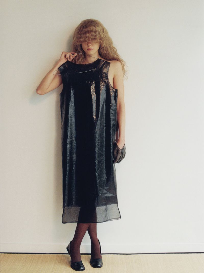 Model with long curly hair wearing a glossy black dress standing against a plain white wall.