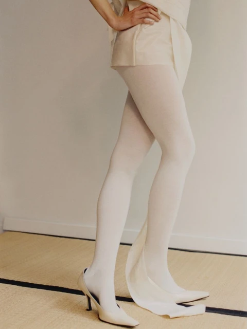 Fashionable white ensemble featuring an adult with tights and heeled shoes, complemented by a neutral studio backdrop and a woven mat.