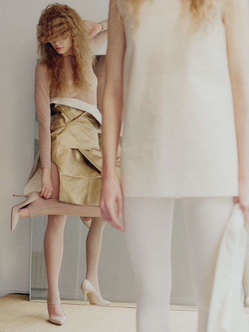 Two women are photographed indoors wearing light neutral-toned clothing, including a gold satin skirt and white top with leggings.