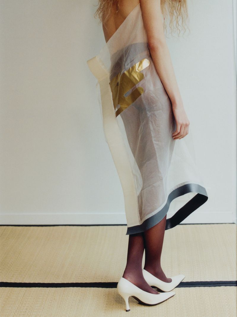 Woman poses indoors wearing a sheer dress with gold detail and white heels, standing on a tan woven rug in a fashion photography style.