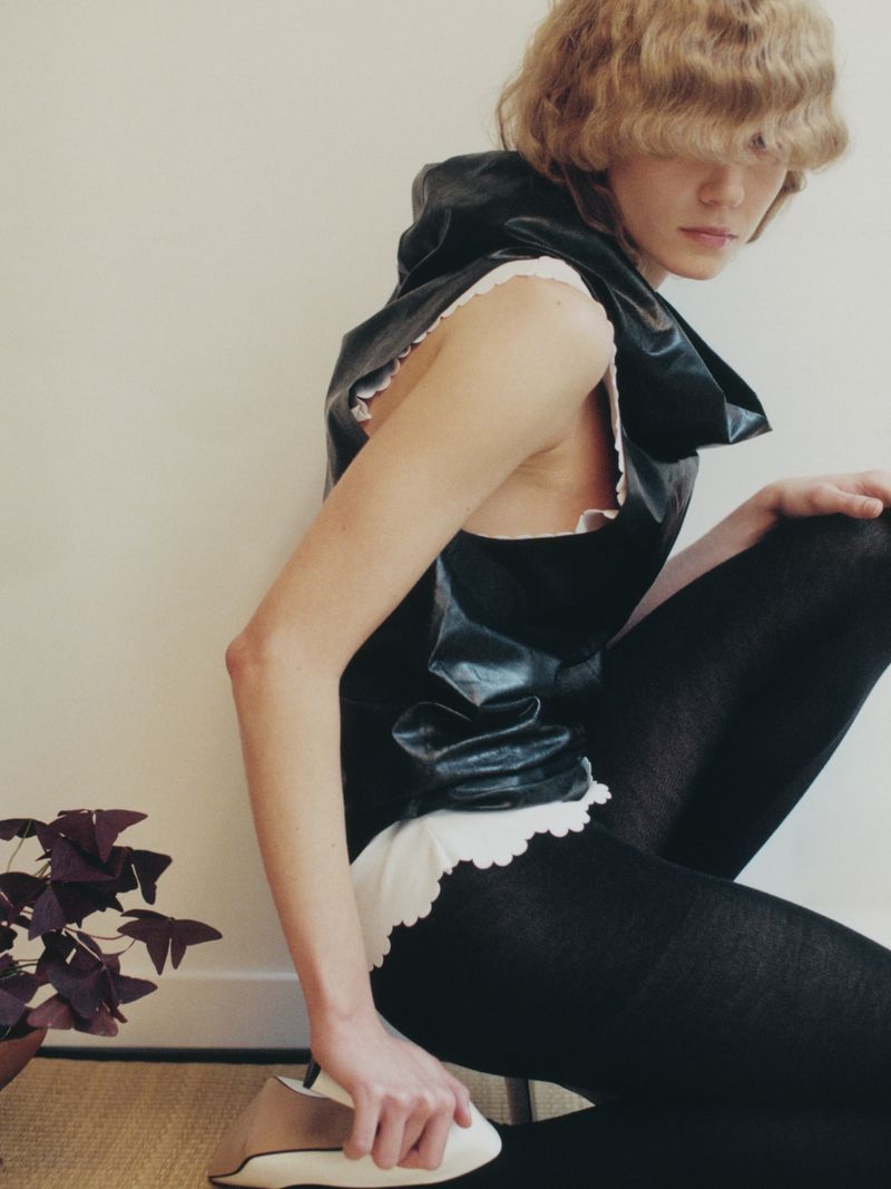 A woman with wavy blonde hair wearing a hooded black top and black tights kneels on the floor with a plant nearby.