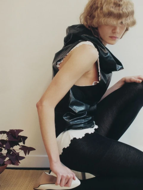 A woman with wavy blonde hair wearing a hooded black top and black tights kneels on the floor with a plant nearby.