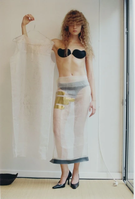 A stylish woman poses indoors wearing a sheer skirt, bra cups, and heels while holding a sheer garment on a hanger in her hand.