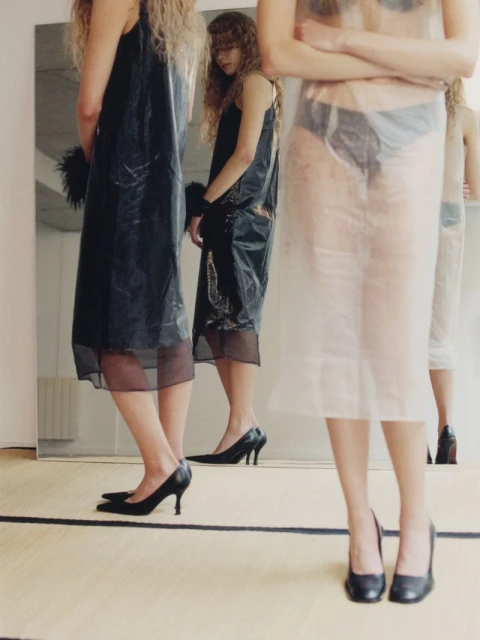 Three fashion models in semi-transparent and black slip dresses and heels pose in a minimalist photo studio reflected in a mirror.