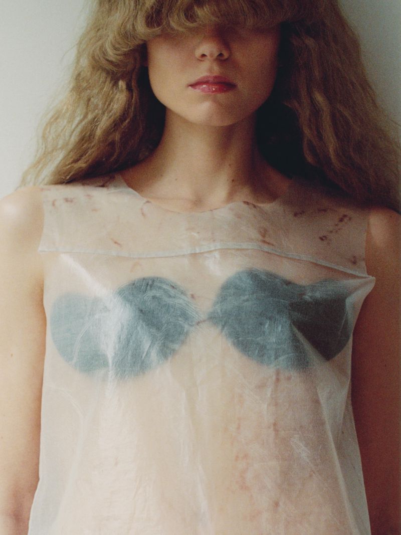 Portrait of a young woman with curly blonde hair partially obscuring her face, wearing a translucent top and bra against a white background.
