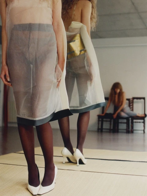 Indoor shot featuring two partially transparent white dresses on models, one reflected in a mirror with a seated woman in background.