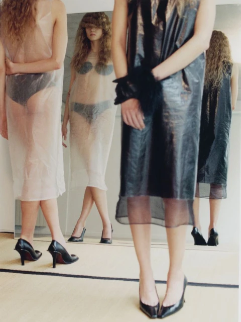 Three women in sheer dresses and heels stand near a mirror, reflecting the scene in a minimalist indoor setting.