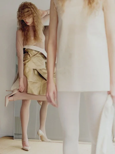 Two female models posing indoors with one model standing in the foreground and another reflected in a mirror behind her.