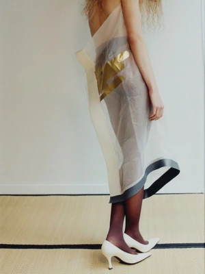 Model wearing a sheer paneled dress and white heels against a neutral wall