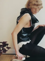 Model with blonde hair covering eyes wearing a black hooded vest and dark tights crouching indoors