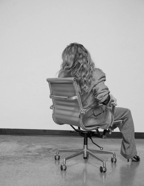 Black and white shot of a woman with curly hair wearing a suit, sitting in a rolling chair, against a stark white wall backdrop.
