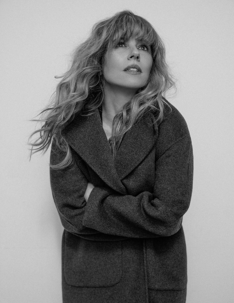 Black-and-white portrait of a blonde woman wearing a wool coat and looking up, captured in a studio setting with even lighting.