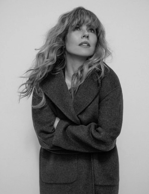 Black-and-white portrait of a blonde woman wearing a wool coat and looking up, captured in a studio setting with even lighting.