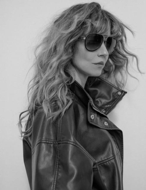 Black and white portrait of a woman with curly hair wearing sunglasses and a leather jacket against a white background.