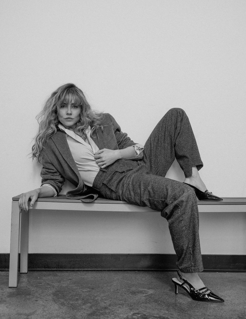 A blonde woman in a pantsuit reclines on a bench against a white wall, gazing into the camera in a black and white studio shot.