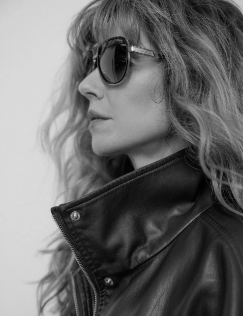 Black and white portrait of a woman with wavy hair, wearing sunglasses and a leather jacket, against a plain background, showcasing fashion.