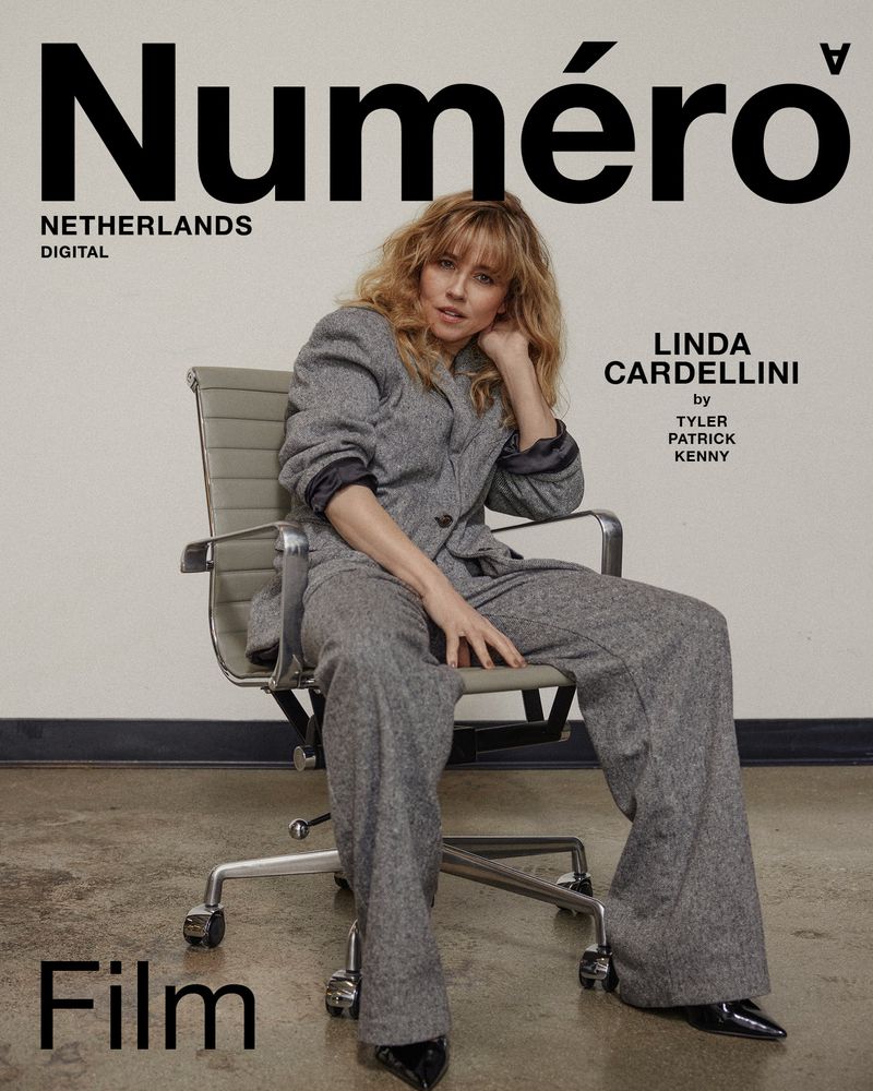 Linda Cardellini poses on an office chair for a Numéro Netherlands magazine cover, wearing a grey suit and black heels, with minimal makeup.