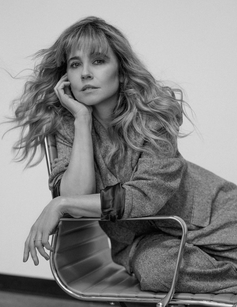 Black and white portrait of a seated woman with long, wavy hair, wearing a herringbone suit, in a photo studio with soft lighting.
