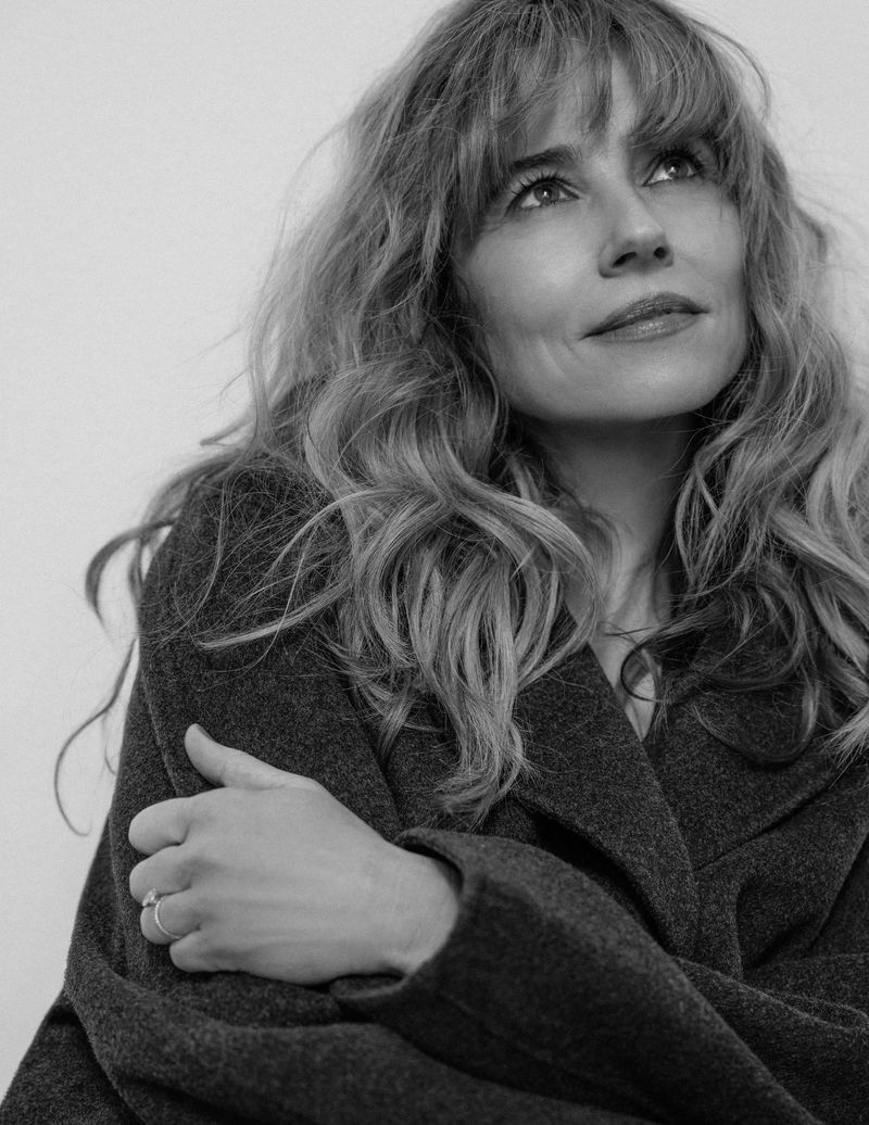 Black and white portrait of a fair-skinned adult woman with long blonde wavy hair wearing a wool coat with her arms crossed.