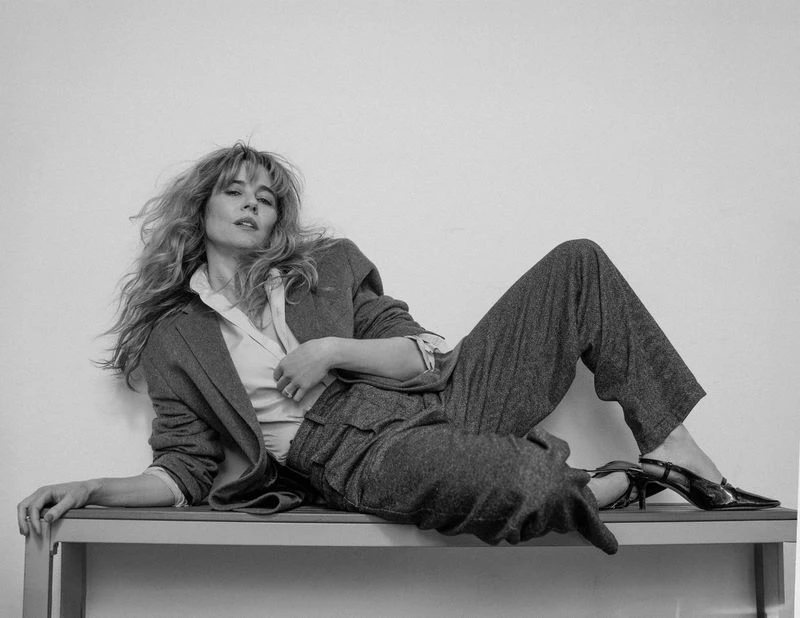 Black and white portrait of an adult woman with long blonde hair, wearing a blazer and pants, reclining on a surface indoors.