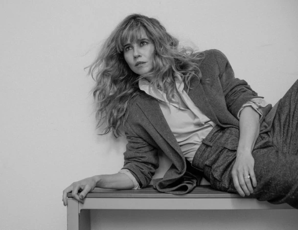 Black and white portrait of Linda Cardellini reclining on a bench wearing a tailored blazer and trousers for Numero Netherlands.