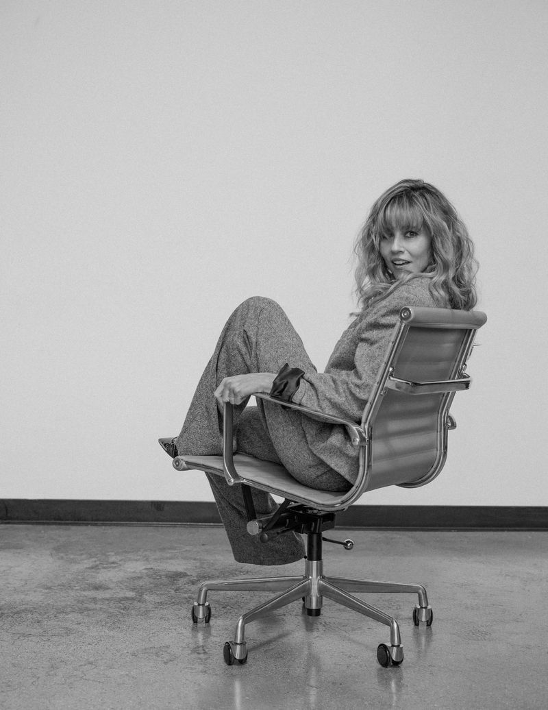 Monochrome portrait of a woman in a suit sitting in an office chair, facing the camera with a relaxed expression, against a plain wall.