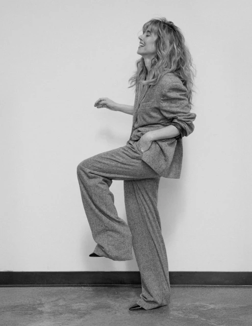 Monochrome portrait of a woman with long, wavy hair in a gray suit, leg raised in a playful pose against a minimalist background.