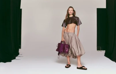 Model Natasha Poly wearing a dark cropped top and grey skirt holding a purple handbag against a white studio background.