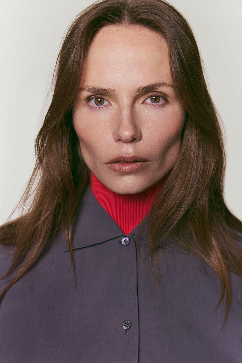 Headshot of a woman with long brown hair wearing a grey button-down shirt and red turtleneck against a neutral studio background.