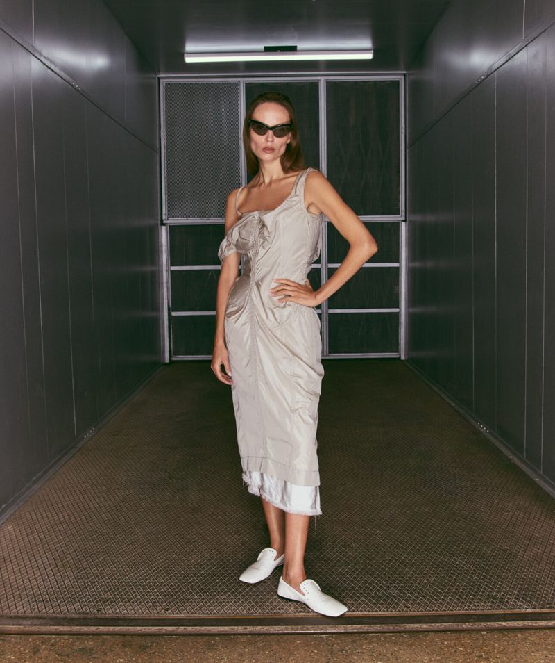 Elegant adult female posing indoors wearing a draped grey dress with white flats, and sunglasses, set against a gray corridor backdrop.