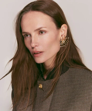 Close-up shot of a woman with long brown hair wearing a houndstooth jacket and large gold and pearl earrings, posing indoors.
