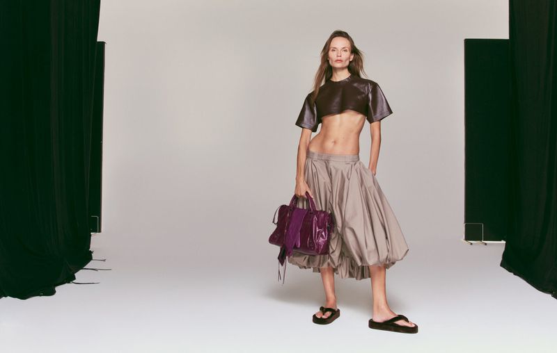 Fashion shot of a woman in a brown crop top and grey skirt holding a purple bag, standing in a photo studio against a white background.