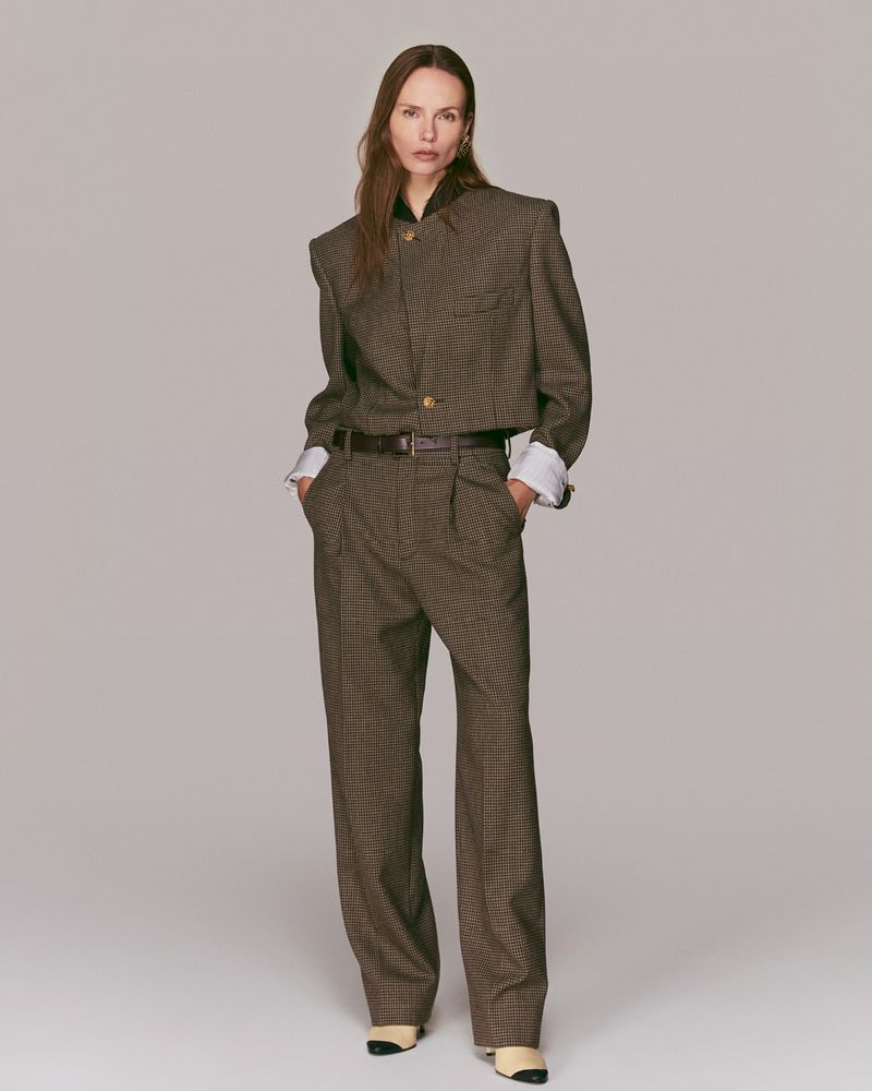 An adult female fashion model in a brown patterned suit stands in a neutral studio with soft lighting and minimal background details.