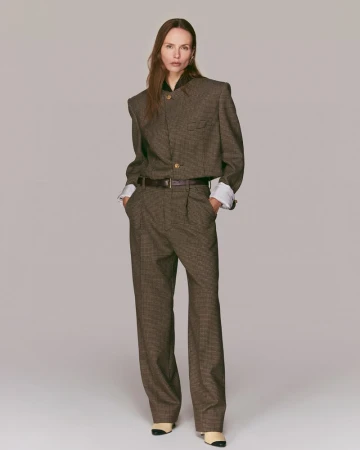 An adult female fashion model in a brown patterned suit stands in a neutral studio with soft lighting and minimal background details.