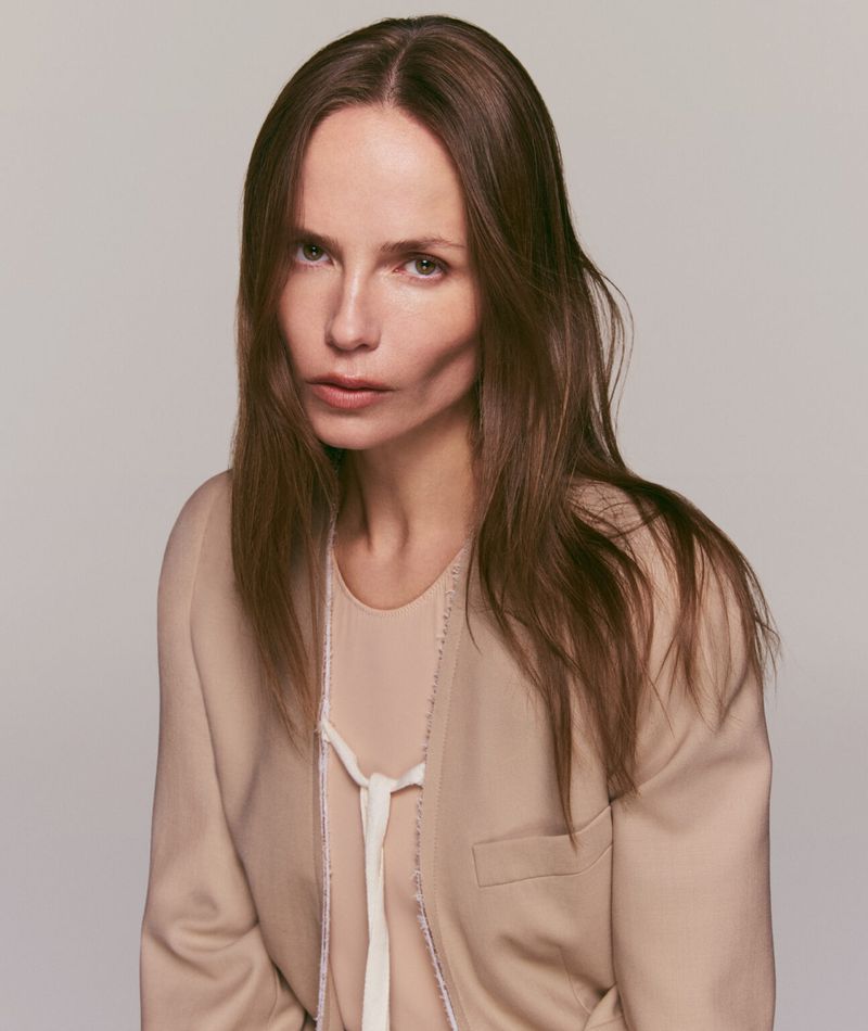Portrait of an adult woman with long brown hair looking at the camera, wearing a beige blazer and a light pink blouse in a photo studio.